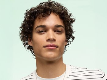 Garnier Hair Care model with short, tousled dark curly hair styled naturally, wearing a white and black striped shirt, posed against a mint green background for a men's grooming lifestyle shot