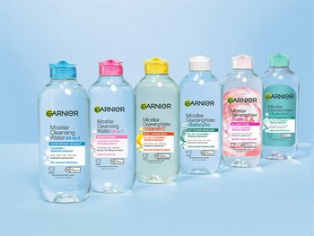 A row of six different Garnier Micellar Cleansing Water bottles, each with a different colored cap (blue, pink, light green, light blue, peach, light green again) and variations in formula, on a light blue surface.