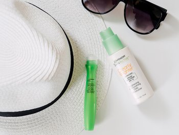 A white straw hat, sunglasses, a green Garnier SkinActive Clearly Brighter Anti-Puff Eye Roller, and a green Garnier SkinActive Clearly Brighter Brightening & Smoothing Daily Moisturizer with SPF 15, arranged on a white surface.