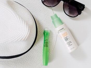 A white straw hat, sunglasses, a green Garnier SkinActive Clearly Brighter Anti-Puff Eye Roller, and a green Garnier SkinActive Clearly Brighter Brightening & Smoothing Daily Moisturizer with SPF 15, arranged on a white surface.