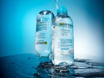 Two bottles of Garnier Micellar Cleansing Waterproof Makeup Remover on a wet surface with splashing water, against a blue background.