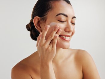 A smiling woman with dark hair pulled back, gently applying a white cream to her cheek.