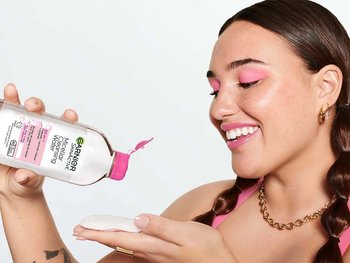 A woman with pink eye makeup and braided pigtails pours Garnier Micellar Cleansing Water (pink cap) onto a cotton pad.