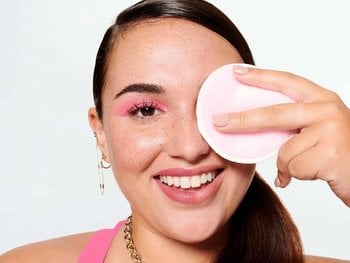 A smiling woman with pink eye makeup uses a pink reusable makeup remover pad to gently clean around her eye.
