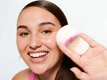 A smiling woman with pink eye makeup holds a makeup removal pad with pink, brown, and beige makeup residue, showing the effectiveness of makeup removal.
