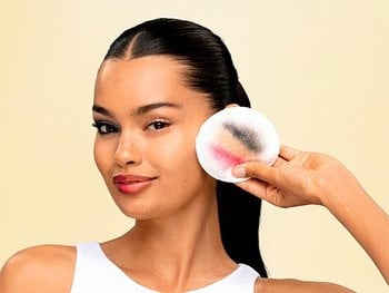 Garnier Micellar Water demonstration showing a woman with sleek dark hair pulled back, glowing skin, and pink lipstick holding a white cotton pad stained with black eye makeup and pink lipstick residue beside her clean cheek.