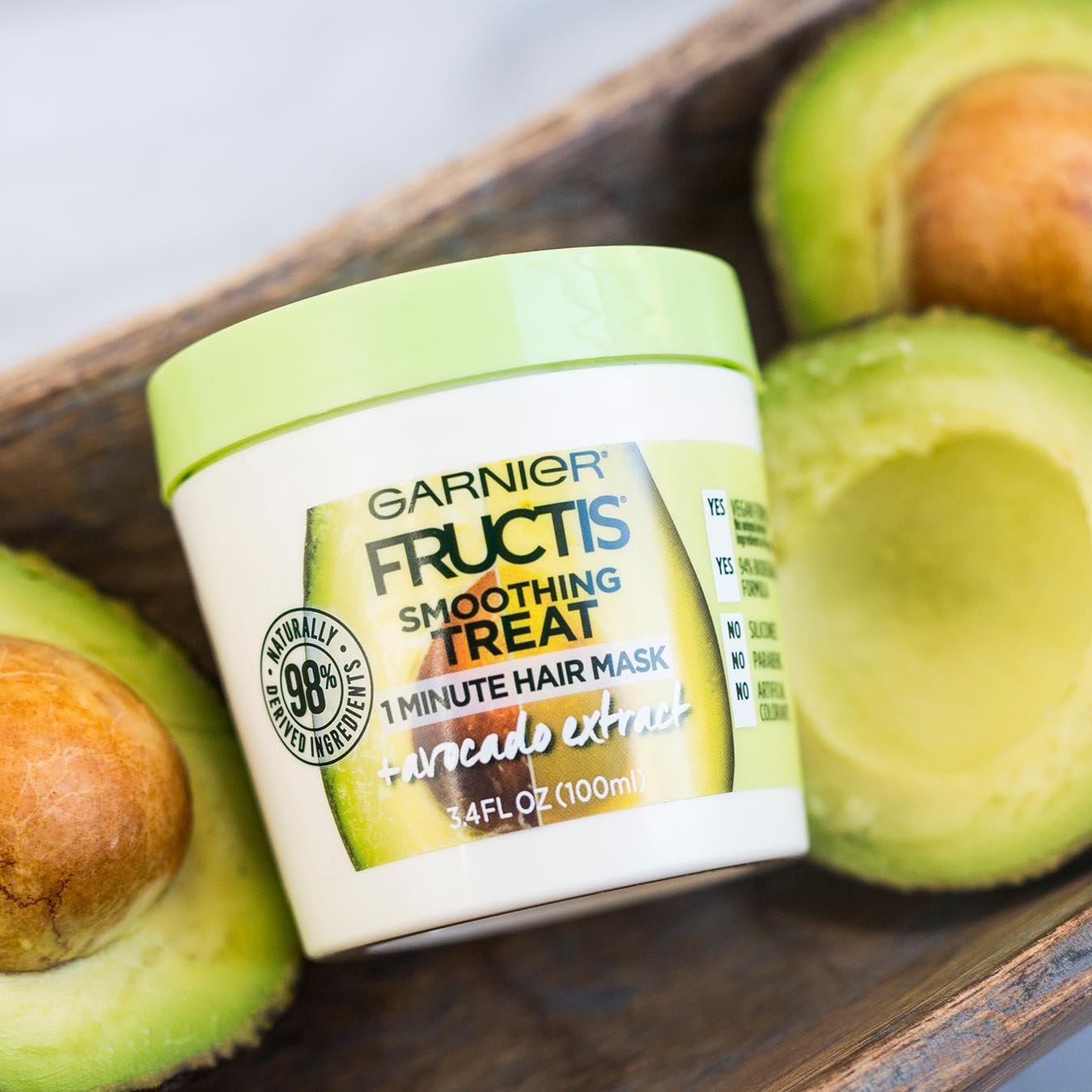 Garnier Fructis Smoothing Treat 1 Minute Hair Mask with Avocado Extract in a wooden trough next to halved avocados on a white marble background.