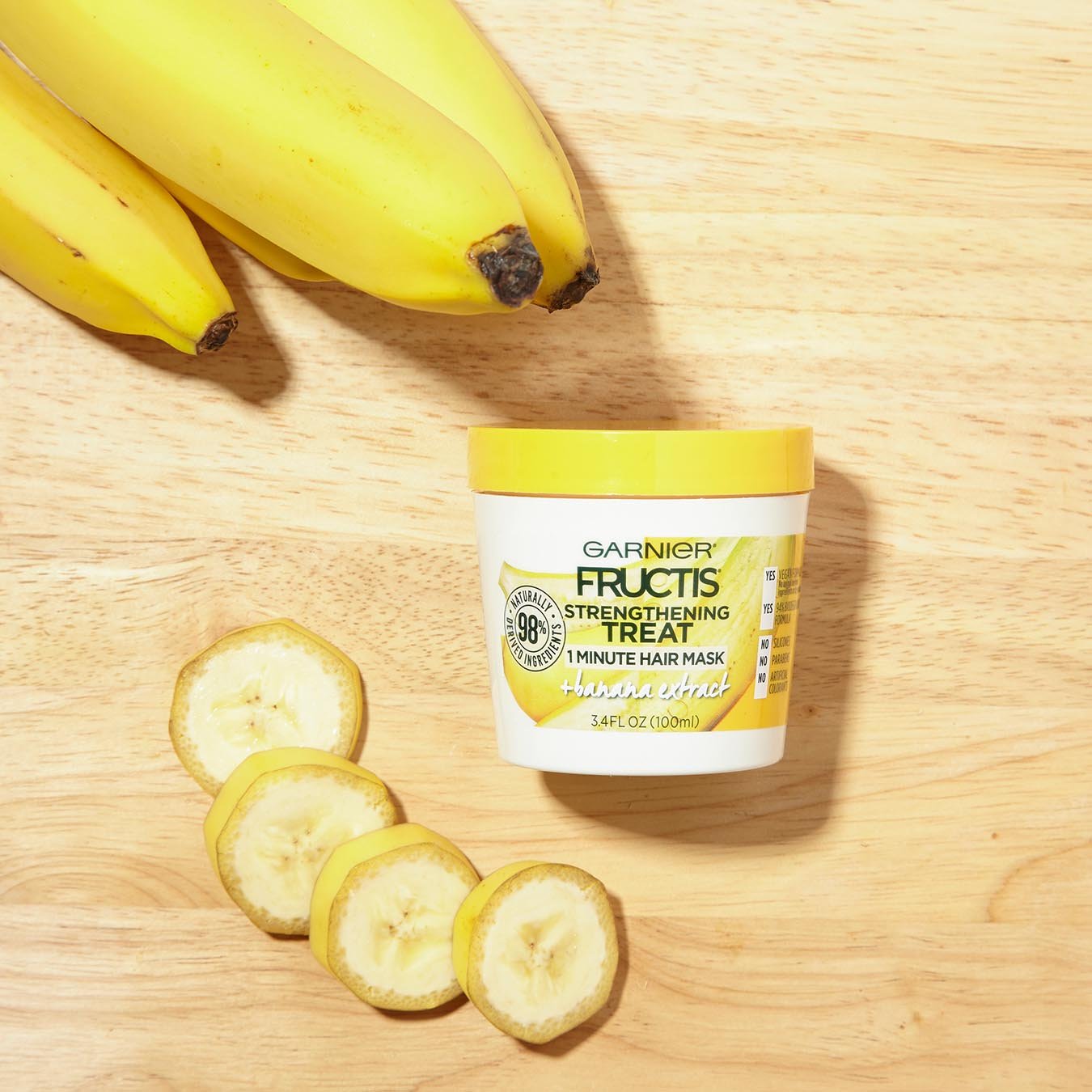 Garnier Fructis Strengthening Treat 1 Minute Hair Mask with Banana Extract on a wooden background next to a bunch of bananas and banana slices.