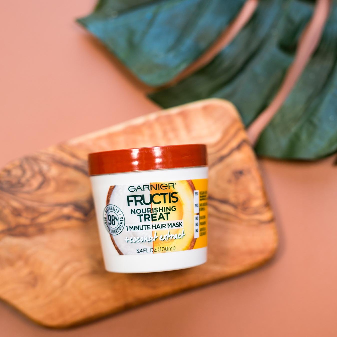 Garnier Fructis Nourishing Treat 1 Minute Hair Mask with Coconut Extract on a wooden cutting board beside a palm frond on a muted red background.