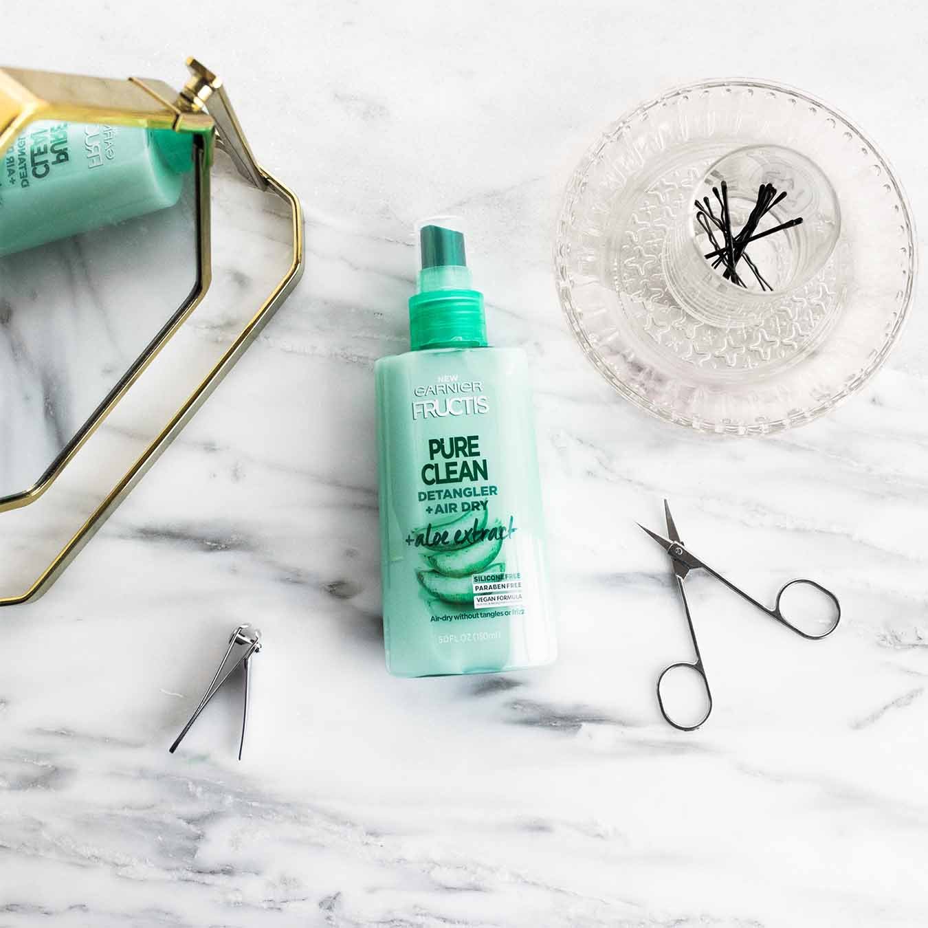 Garnier Fructis Pure Clean Detangler with Aloe Extract reflected in a gold mirror on white marble next to facial hair scissors, fingernail clippers, and a glass cup filled with bobby pins sitting on a glass plate. 