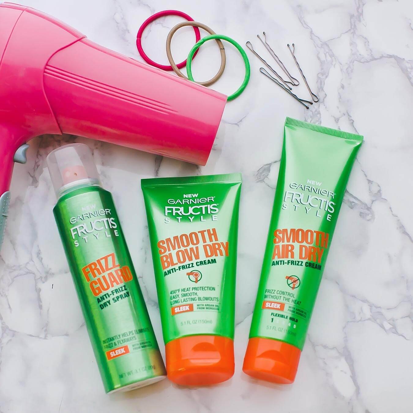 Garnier Fructis Style Frizz Guard Anti-Frizz Dry Spray, Fructis Style Smooth Blow Out Anti-Frizz Cream, and Fructis Style Smooth Air Dry Anti-Frizz Cream on white marble next to a pink blow dryer, four bobby pins, and three hair bands colored pink, brown, and green.