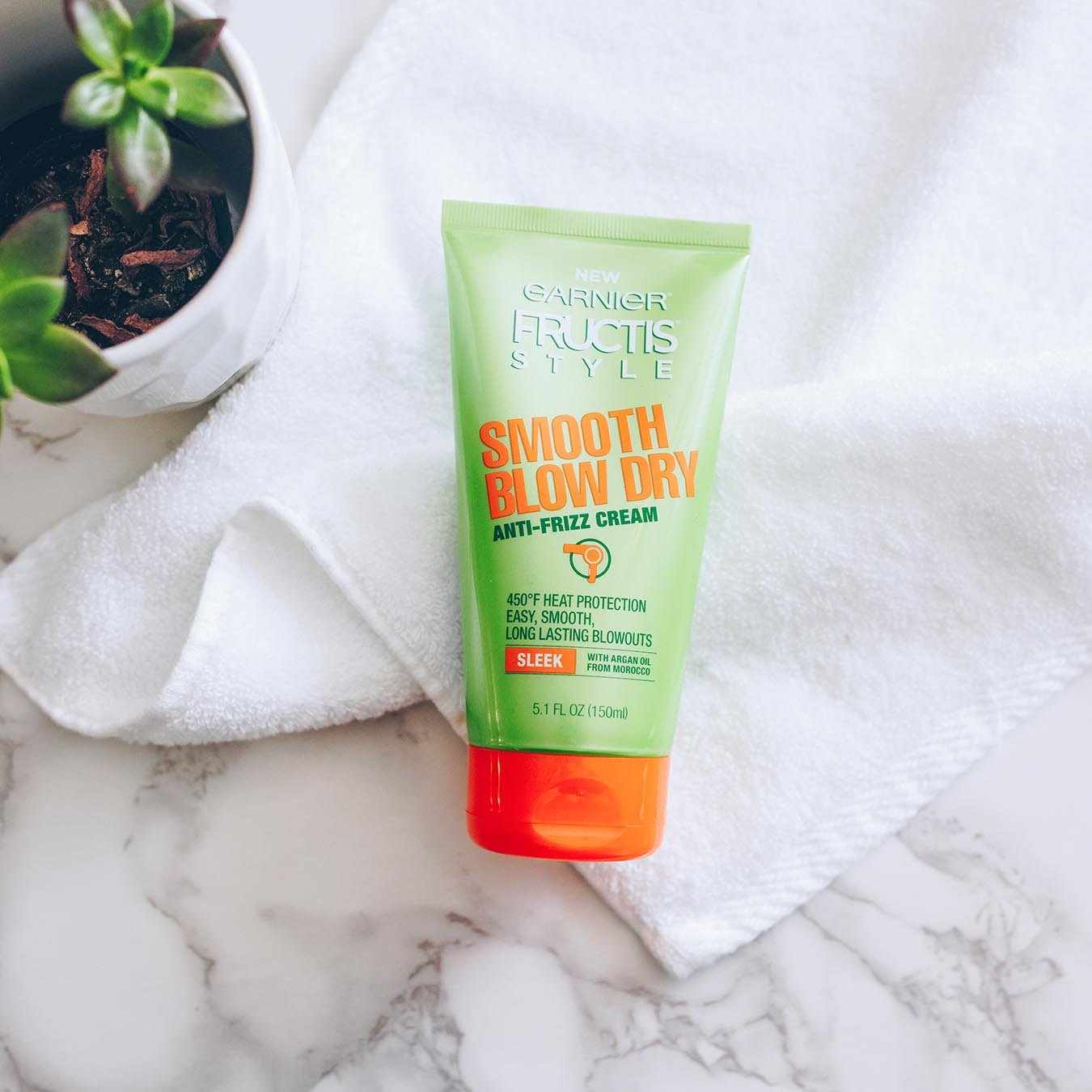 Garnier Fructis Style Smooth Blow Dry Anti-Frizz Cream on a white hand towel on white marble next to a succulent planted in a white faceted pot.