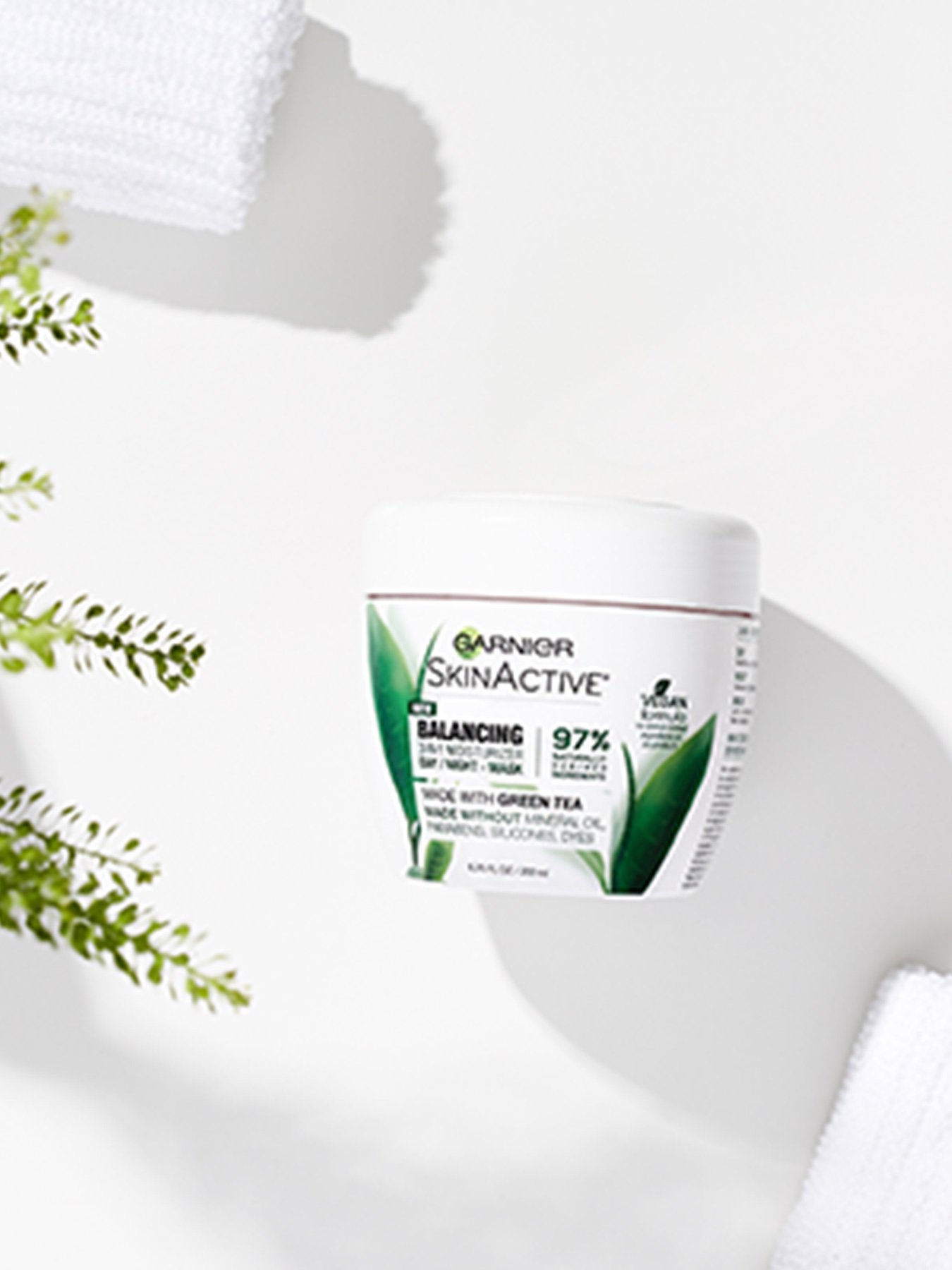 Garnier SkinActive Balancing 3-in-1 Face Moisturizer with Green Tea on a white surface with two white loofahs and some greenery. 