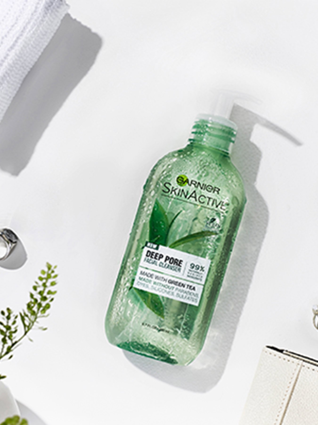 Garnier SkinActive Deep Pore Face Wash with Green Tea on a white surface with a white loofah, white wallet, two silver rings, a white wallet, and a small potted plant.