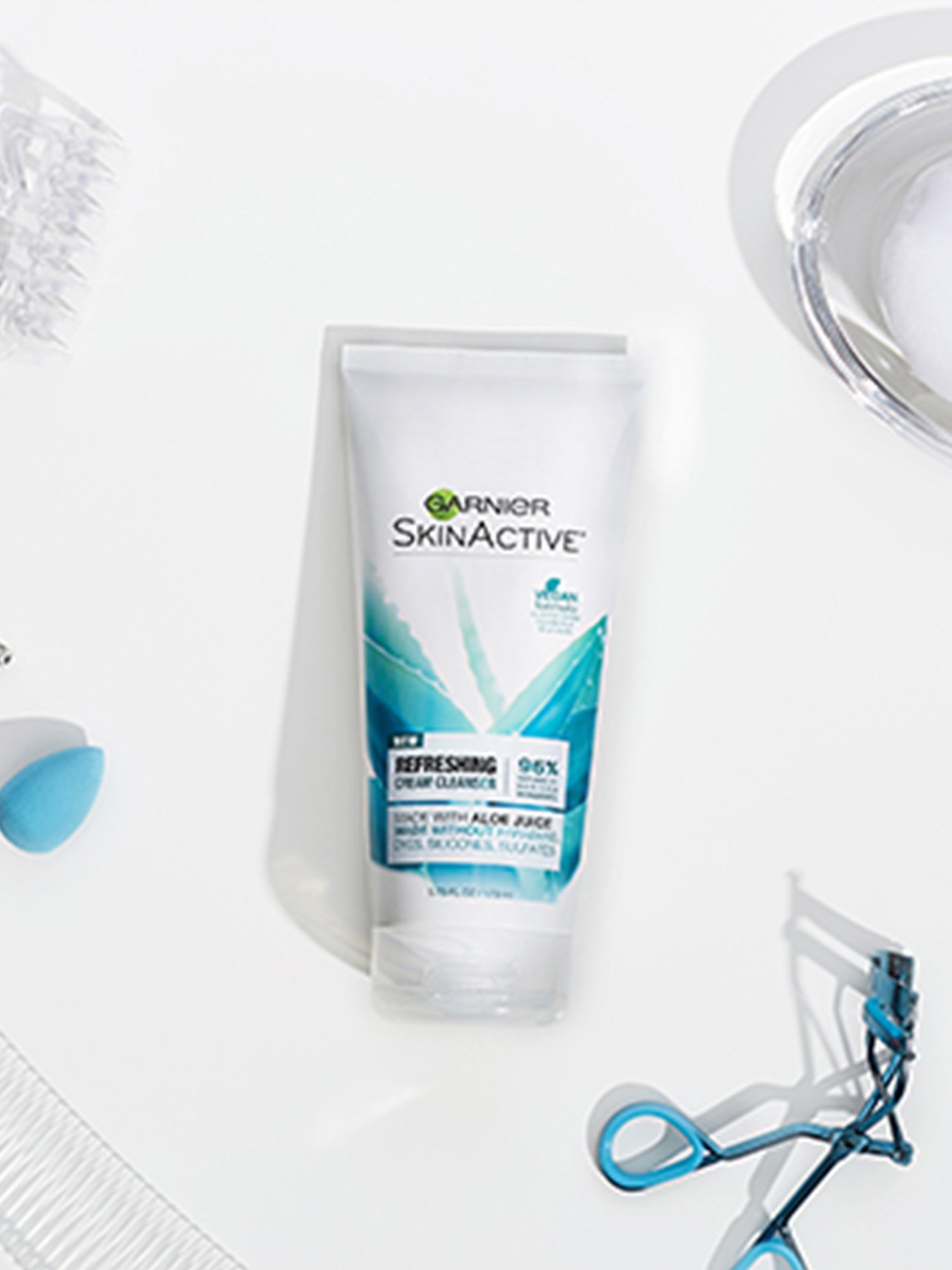 Garnier SkinActive Refreshing Cream Face Wash with Aloe on a white background with a clear glass dish, blue eyebrow crimpers, a clear comb, a clear hair clip, and a blue makeup sponge.