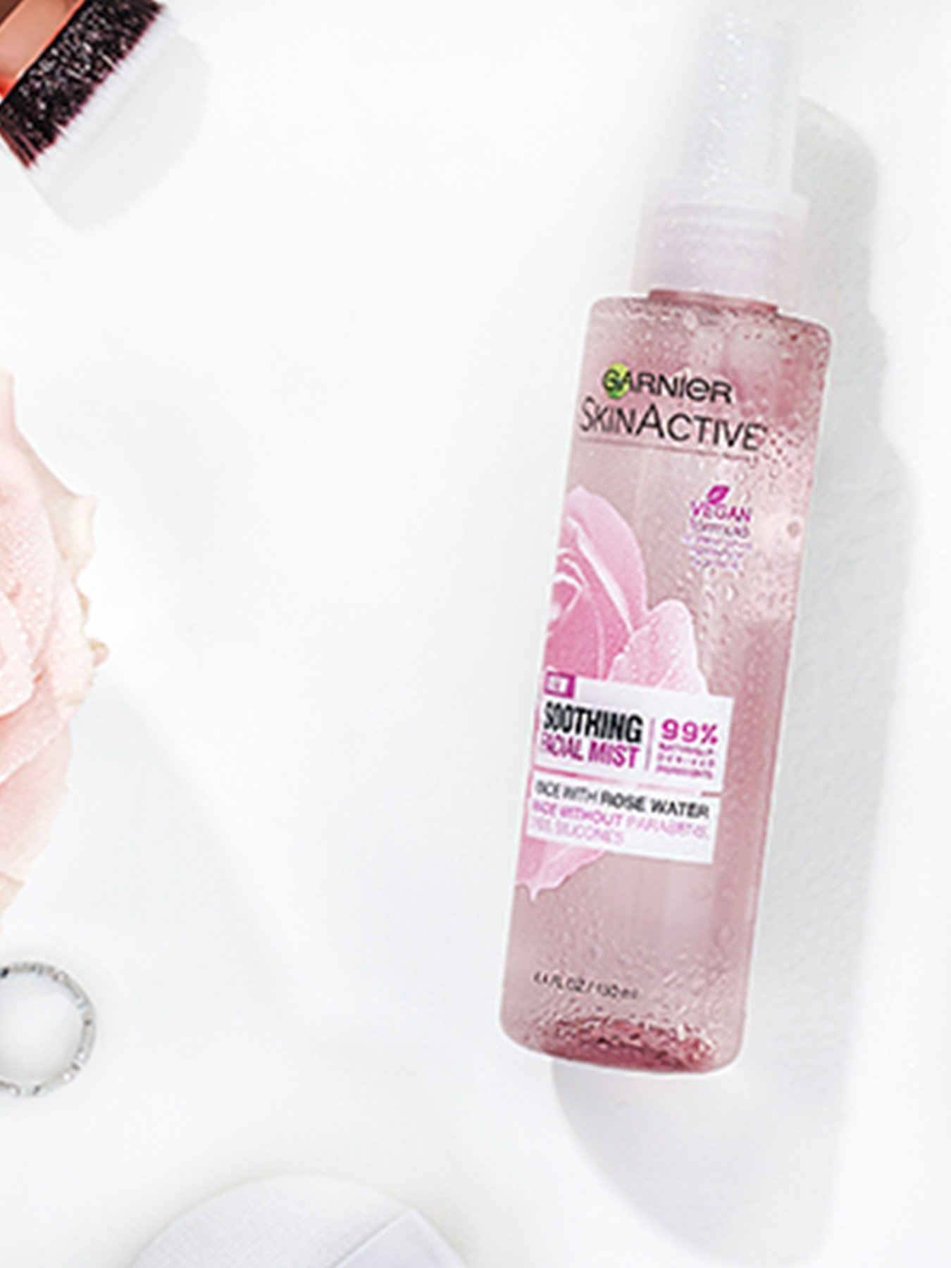 Garnier SkinActive Soothing Facial Mist with Rose Water on a white background with a makeup brush, pink rose, silver ring, and white case.