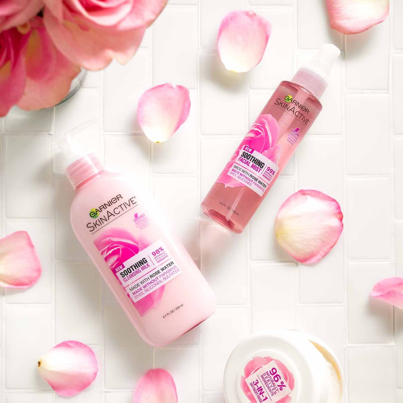 Garnier SkinActive Soothing Cleansing Milk, SkinActive Soothing Facial Mist, SkinActive 3-in-1 Day/Night Mask ajar on white tile with pink rose petals and a vase of pink roses.