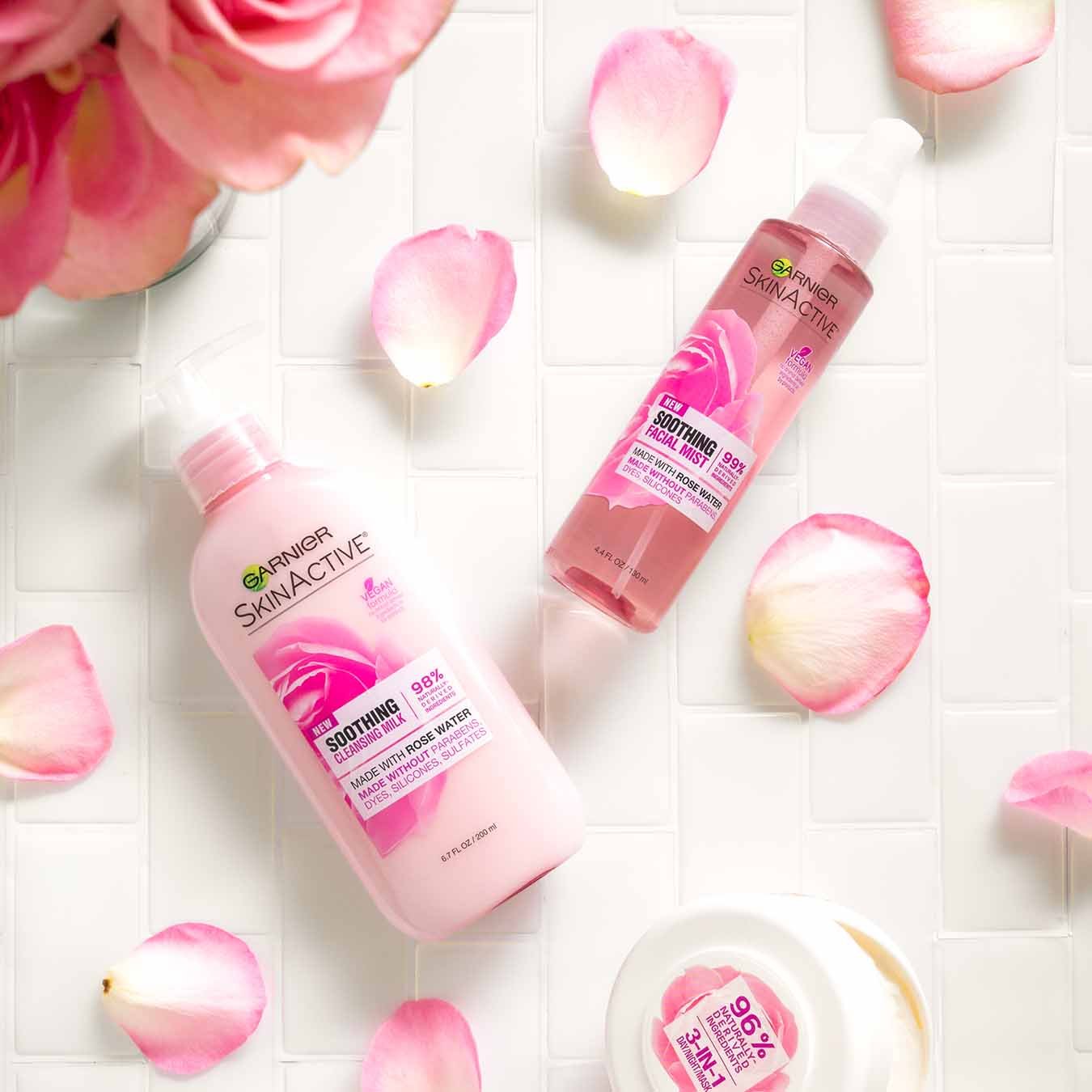 Garnier SkinActive Soothing Cleansing Milk, SkinActive Soothing Facial Mist, SkinActive 3-in-1 Day/Night Mask ajar on white tile with pink rose petals and a vase of pink roses.