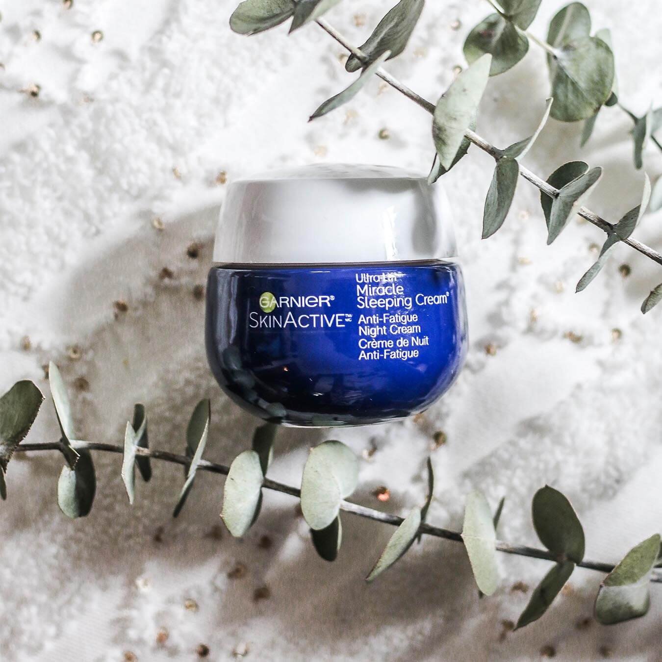 Garnier SkinActive Ultra Lift Miracle Sleeping Cream on a white textured cloth with golden beads with eucalyptus leaves.