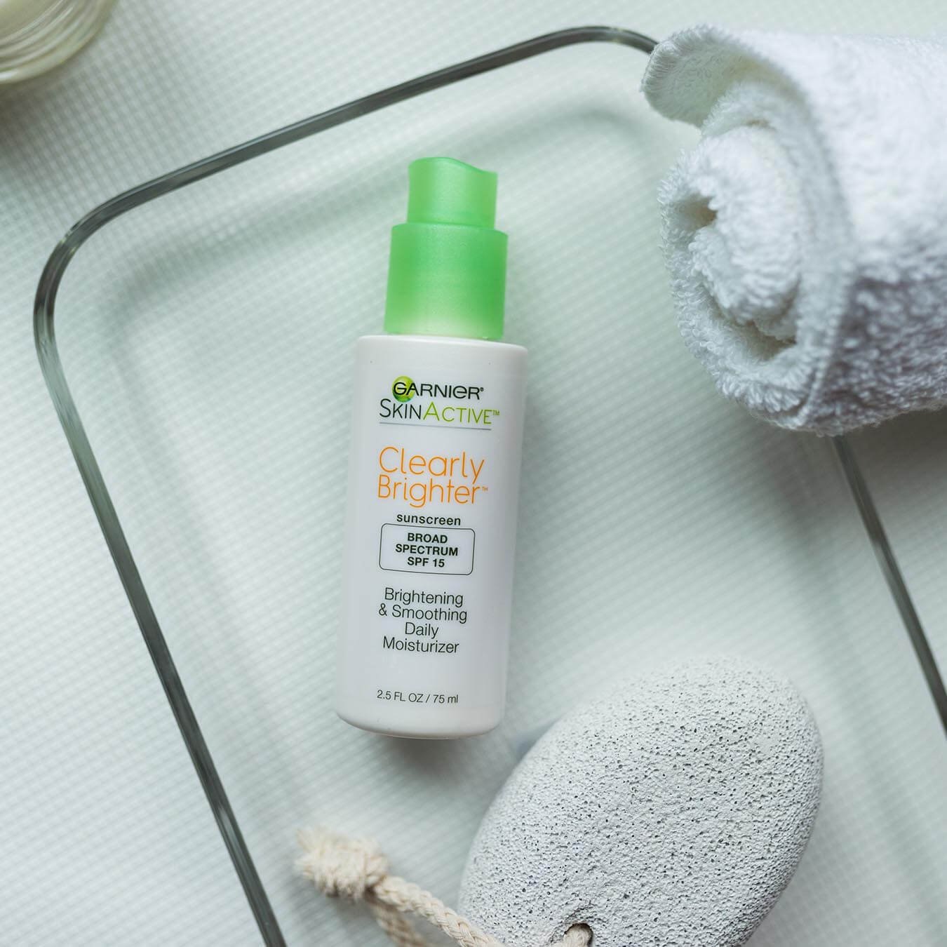 Garnier SkinActive Clearly Brighter Sunscreen Broad Spectrum SPF 15 Brightening & Smoothing Daily Moisturizer sitting in a rectangular glass dish with a pumice stone on a rope on a white mat with a rolled up white hand towel and bowl.