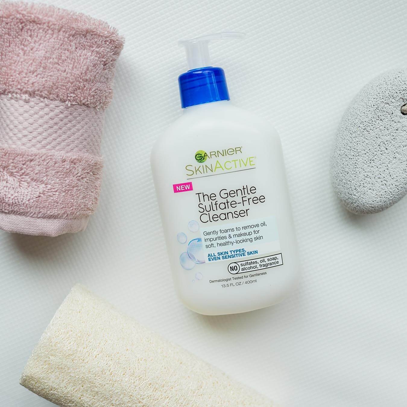 Garnier SkinActive Gentle Sulfate-Free Cleanser on a white mat next to a rolled up pink hand towel, pumice stone on a rope, and loofah.