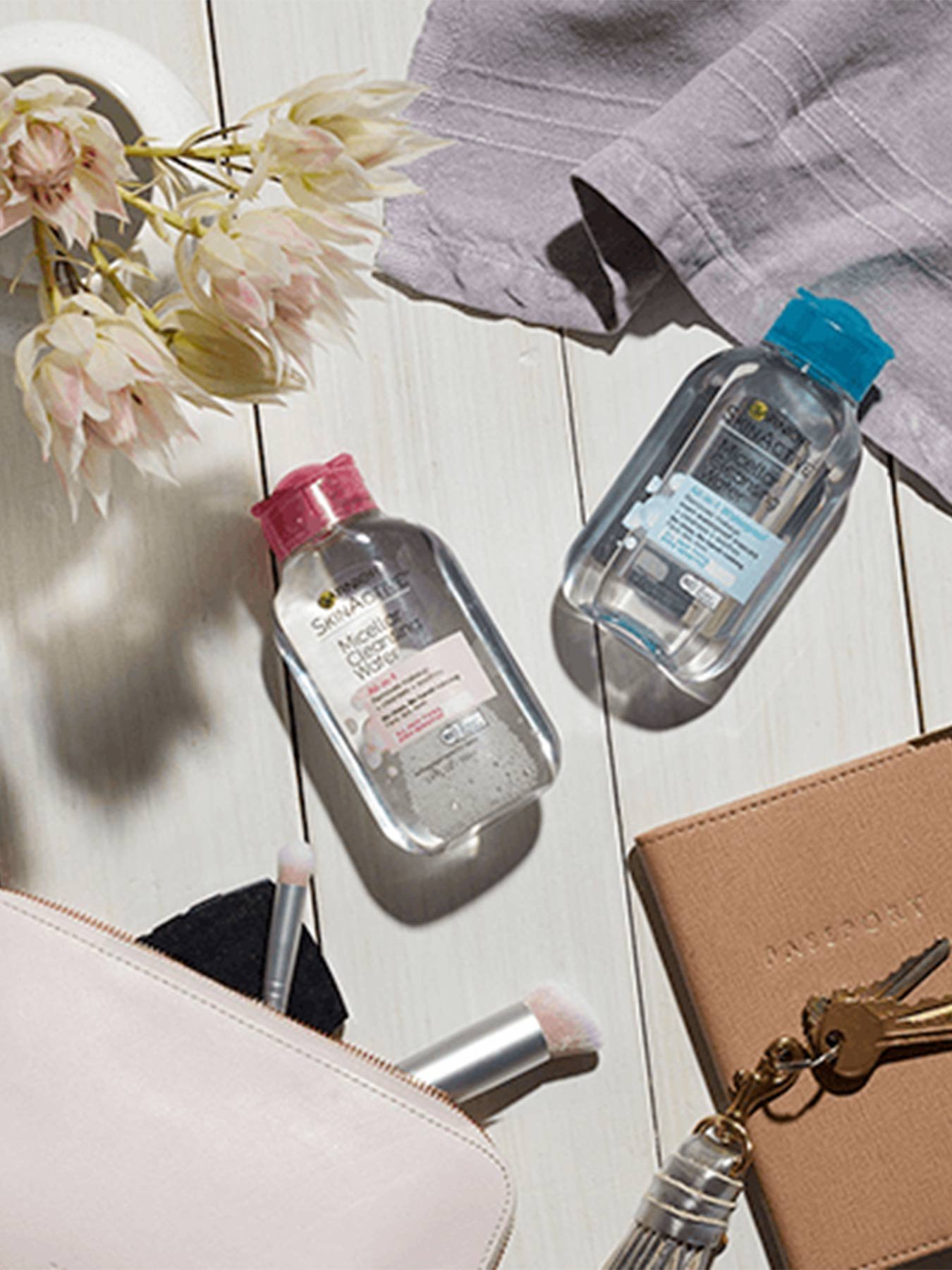A bottle of Micellar Cleansing Water Travel Size and Micellar Waterproof Cleansing Water Travel Size on a white wooden background an open pink makeup bag with makeup brushes sticking out, keys with a gray fob resting on a brown journal, a lavender towel, and a white vase filled with light pink flowers.