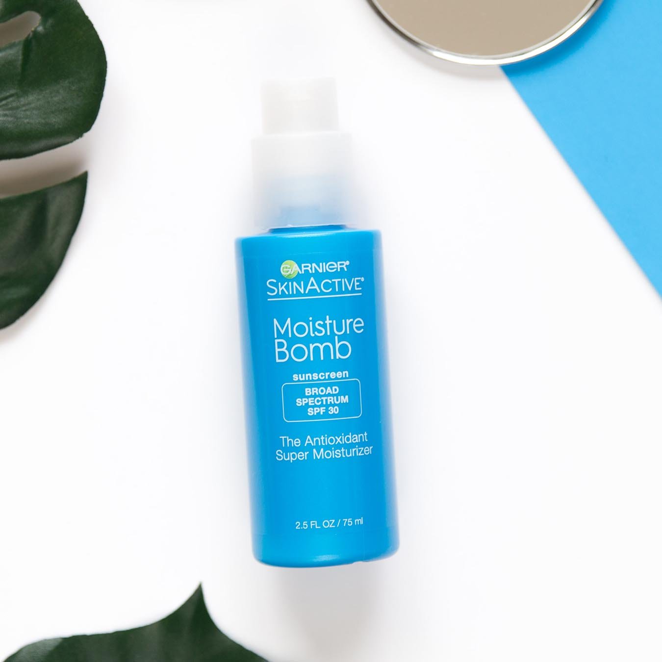 Garnier SkinActive Moisture Bomb Sunscreen Broad Spectrum SPF 30 The Antioxidant Super Moisturizer on a white background next to palm fronds and a mirror partially sitting on a blue piece of paper.