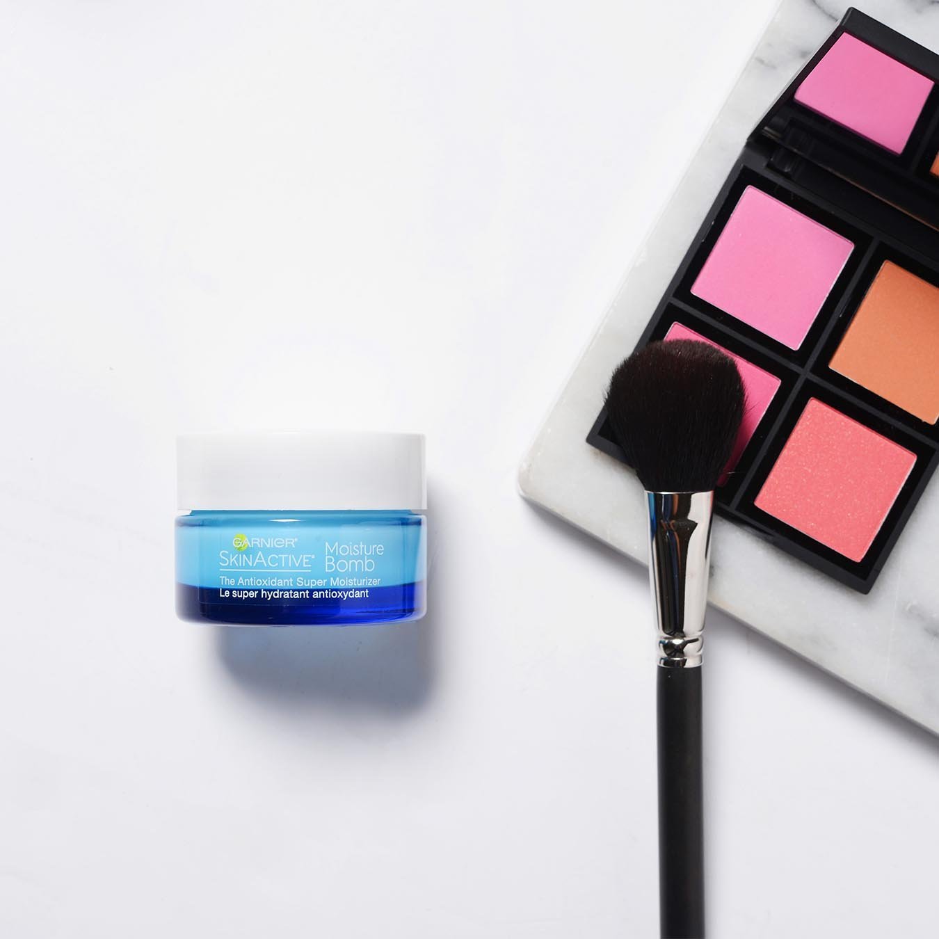 Garnier SkinActive Moisture Bomb The Super Antioxidant Super Moisturizer on a white background next to a palette of pink and orange blushes on a marble slab with a makeup brush.