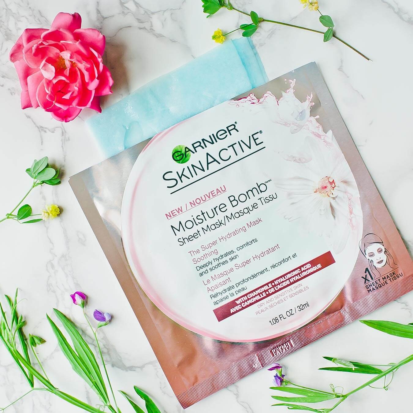 Garnier SkinActive Moisture Bomb Sheet Mask with Chamomile with the blue masks peeking out on white marble next to a pink rose bloom and several wild flowers.