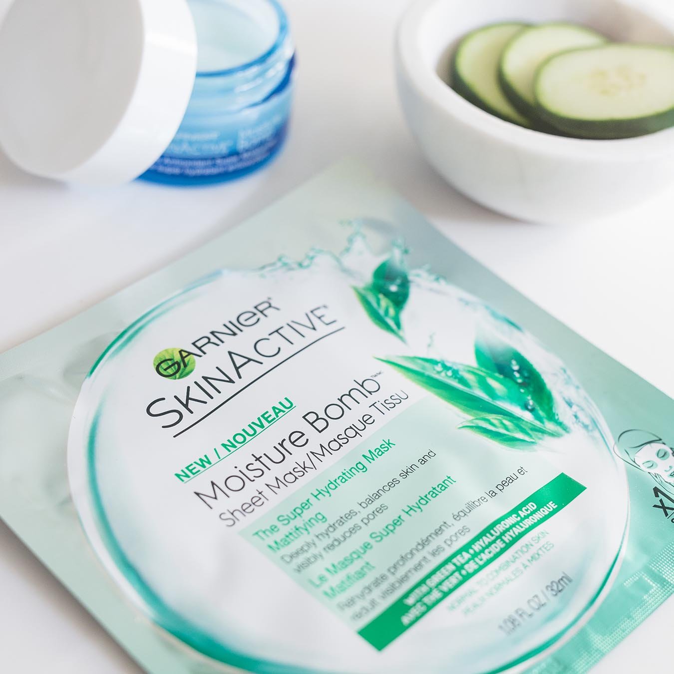 Garnier SkinActive Moisture Bomb Sheet Mask with Green Tea on a white table with SkinActive Antioxidant Super Moisturizer ajar and white marble bowl of sliced cucumbers.