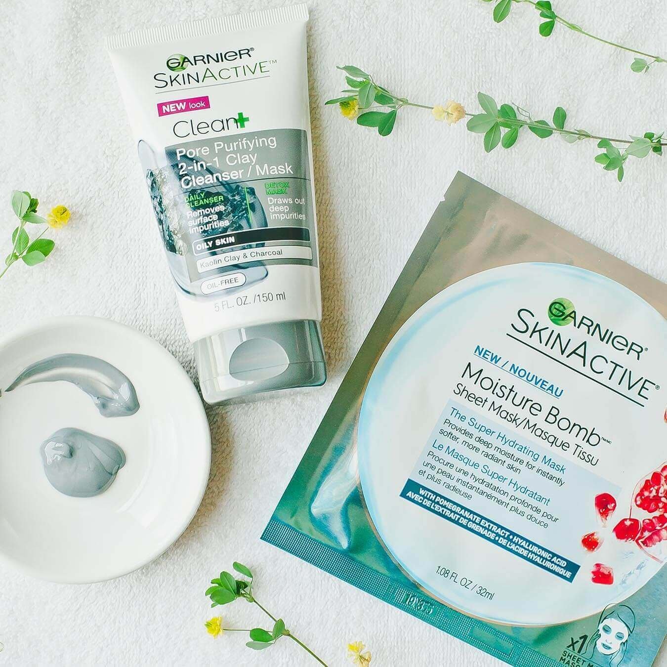 Garnier SkinActive Moisture Bomb Sheet Mask with Pomegranate Extract next to SkinActive Clean+ Pore Purifying 2-in-1 Clay Cleanser/Mask and a white dish with some cleanser/mask squeezed from the tube and smeared in an arc on a white towel strewn with yellow wildflowers.