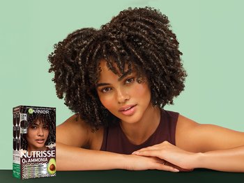 Garnier Nutrisse 0% Ammonia Glossy Curl Color in shade CR4 Glossy Dark Brown: a woman with medium-brown skin and voluminous, defined dark brown coily curls leans on her folded arms on a dark green surface. She wears a burgundy sleeveless top and tilts her head with a soft, slightly parted-lip expression against a mint green background. In the lower left is the dark maroon product box with the Garnier logo, "NUTRISSE 0% AMMONIA," "GLOSSY CURL COLOR," the claims "3x shinier curls, up to 5x less breakage" and "100% undetectable gray coverage," a green avocado graphic, and "Permanent Color, Vegan Formula."