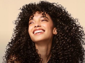 A woman with voluminous, tightly coiled dark brown curly hair tilts her head back, laughing joyfully with her eyes closed against a soft beige background, showcasing full, defined ringlets and glowing skin.
