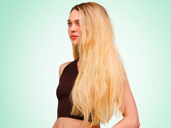 A side-profile lifestyle shot of a woman with light skin and long, straight, slightly frizzy blonde hair wearing a dark brown crop top, posed against a soft mint green gradient background.