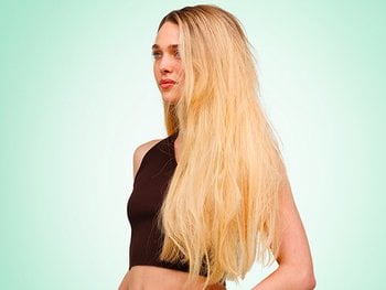 A side-profile lifestyle shot of a woman with light skin and long, straight, slightly frizzy blonde hair wearing a dark brown crop top, posed against a soft mint green gradient background.