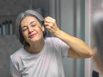 Mature woman applying serum on face in bathroom mirror
