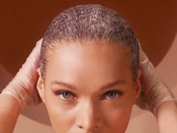 Front portrait shot of model with grey eyes and textured dyed hair. She is wearing gloves as she pulls hair back in the midst of her dyeing process.