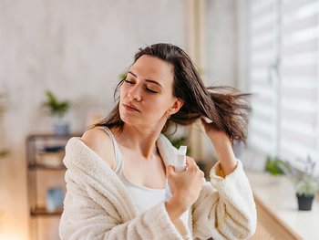 Woman with brown hair applies a spray to her hair with eyes closed.
