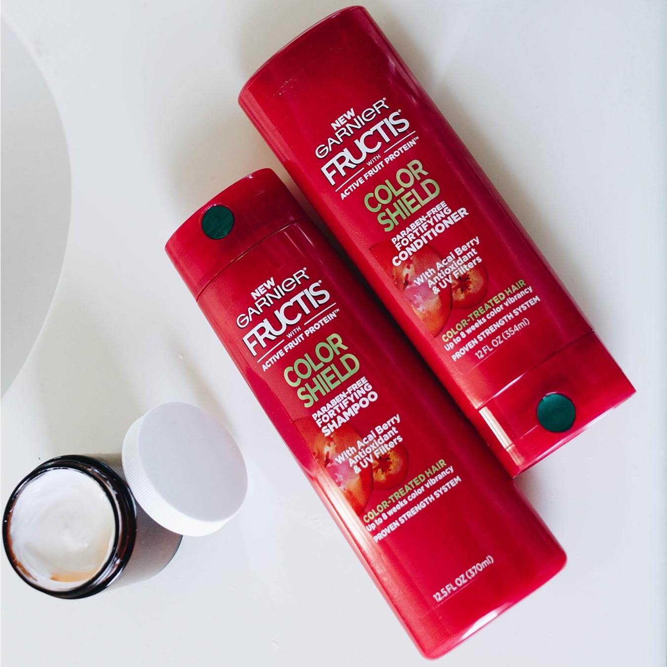 Garnier Fructis Color Shield Fortifying Shampoo and Fructis Color Shield Fortifying Conditioner on a white table next to an open facial cream container.