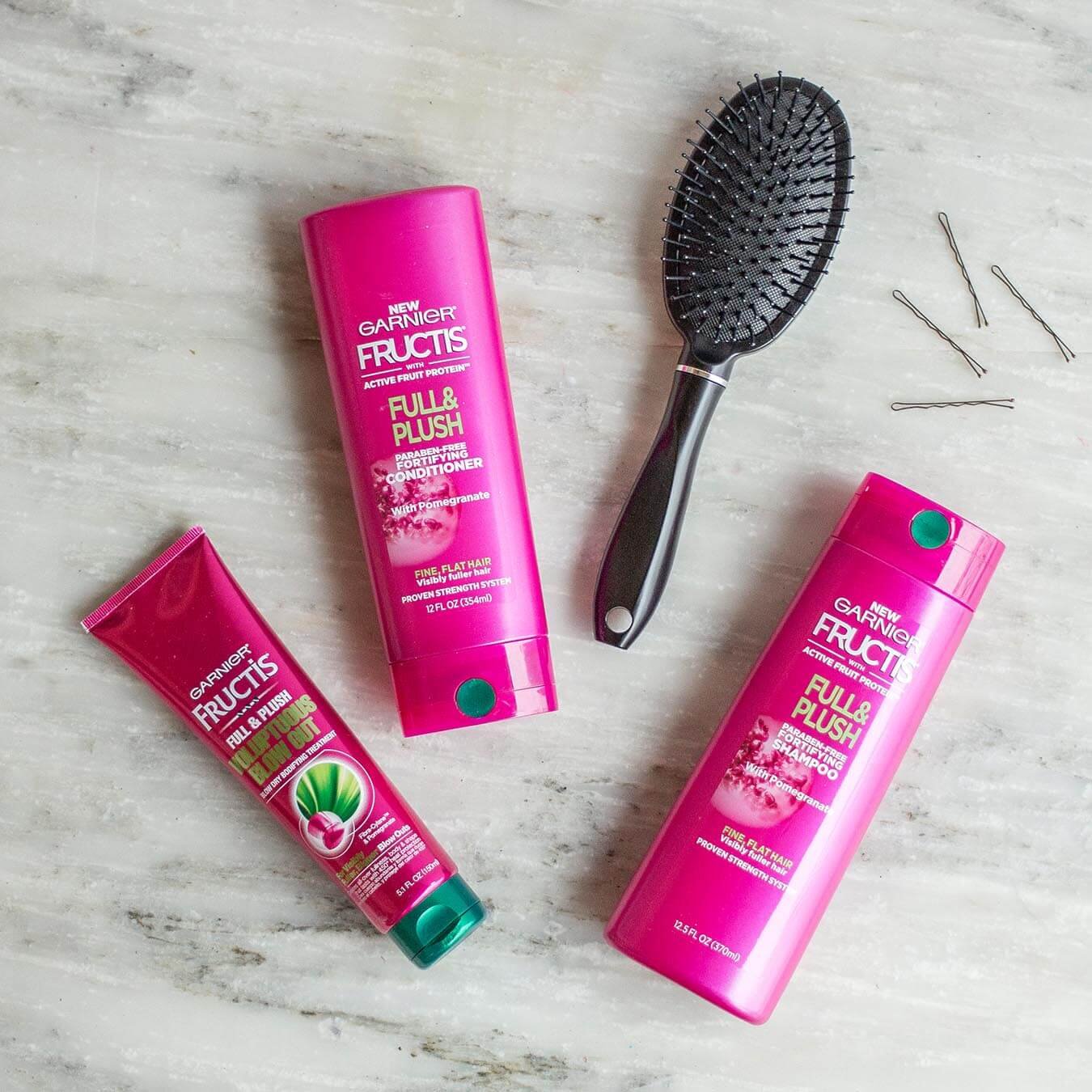 Garnier Full & Plush Shampoo, Full & Plush Conditioner, and Full & Plush Voluptuous Blow Out on gray marble next to four bobby pins and a hair brush.
