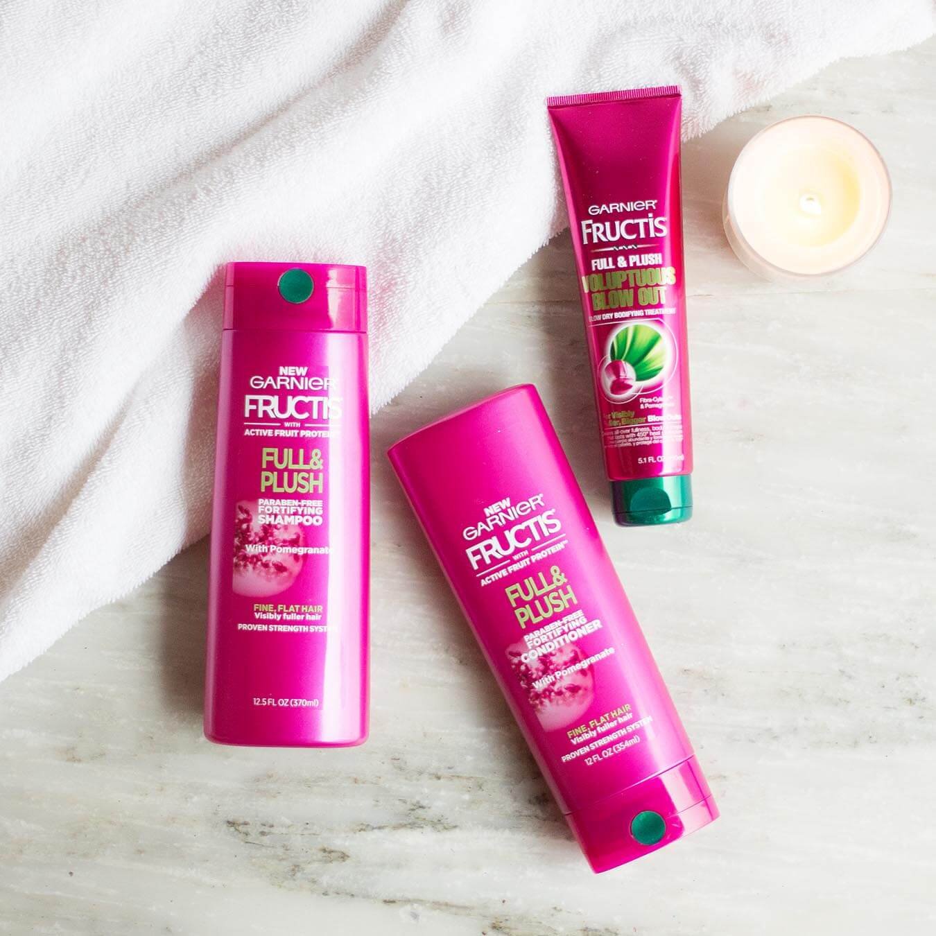 Garnier Full & Plush Shampoo, Full & Plush Conditioner, and Full & Plush Voluptuous Blow Out on gray marble and a white bath towel next to a lit tea candle.