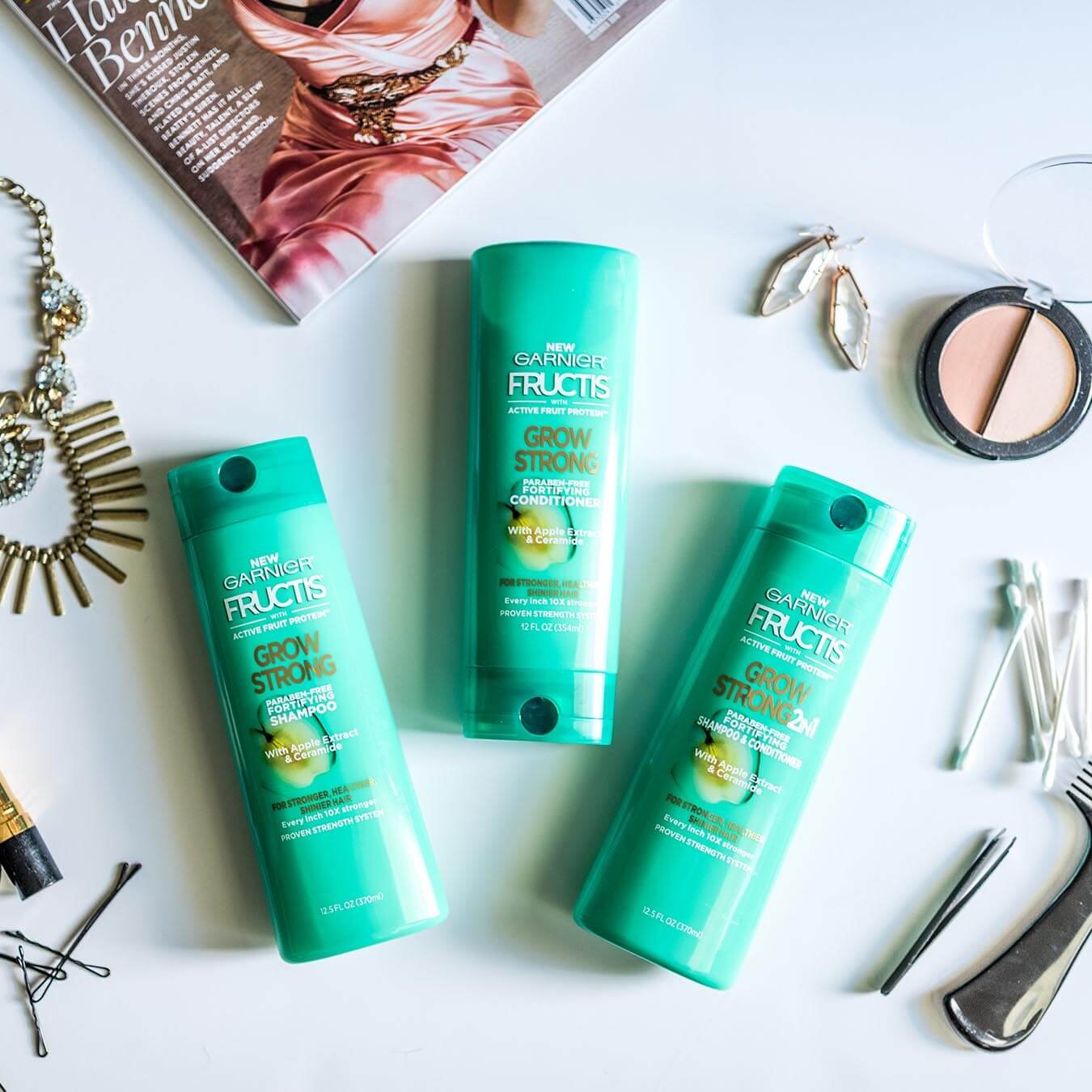 Garnier Fructis Grow Strong Shampoo, Fructis Grow Strong Conditioner, and Fructis Grow Strong 2-in-1 on a blueish-white background next to gold earrings and necklace, bobby pins, tweezers, q-tips, a comb, and two shades of blush.