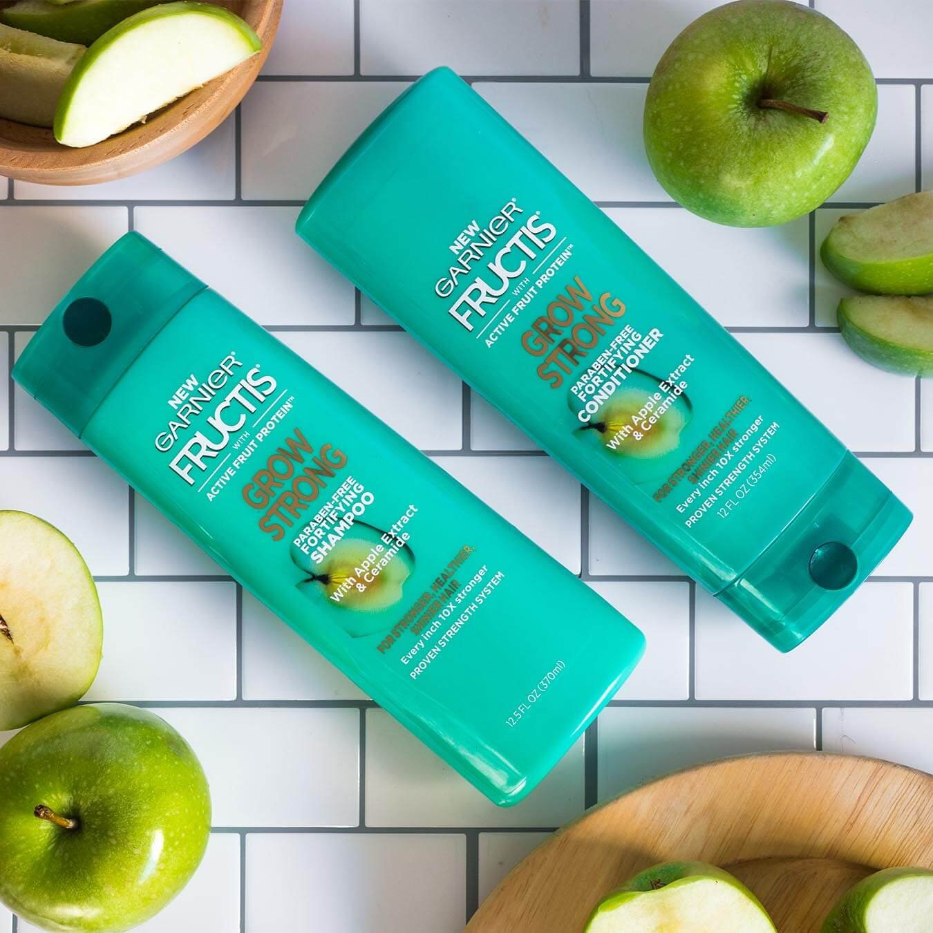 Garnier Fructis Grow Strong Shampoo and Fructis Grow Strong Conditioner on a white tiled background with sliced granny smith apples on wooden plates and in wooden bowls.