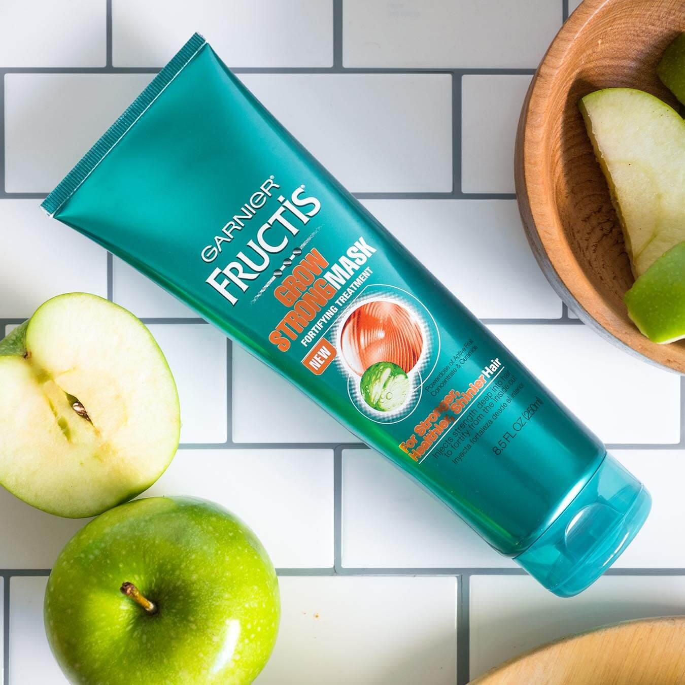 Garnier Fructis Grow Strong mask on a white tiled background with sliced granny smith apples on wooden plates and in wooden bowls.