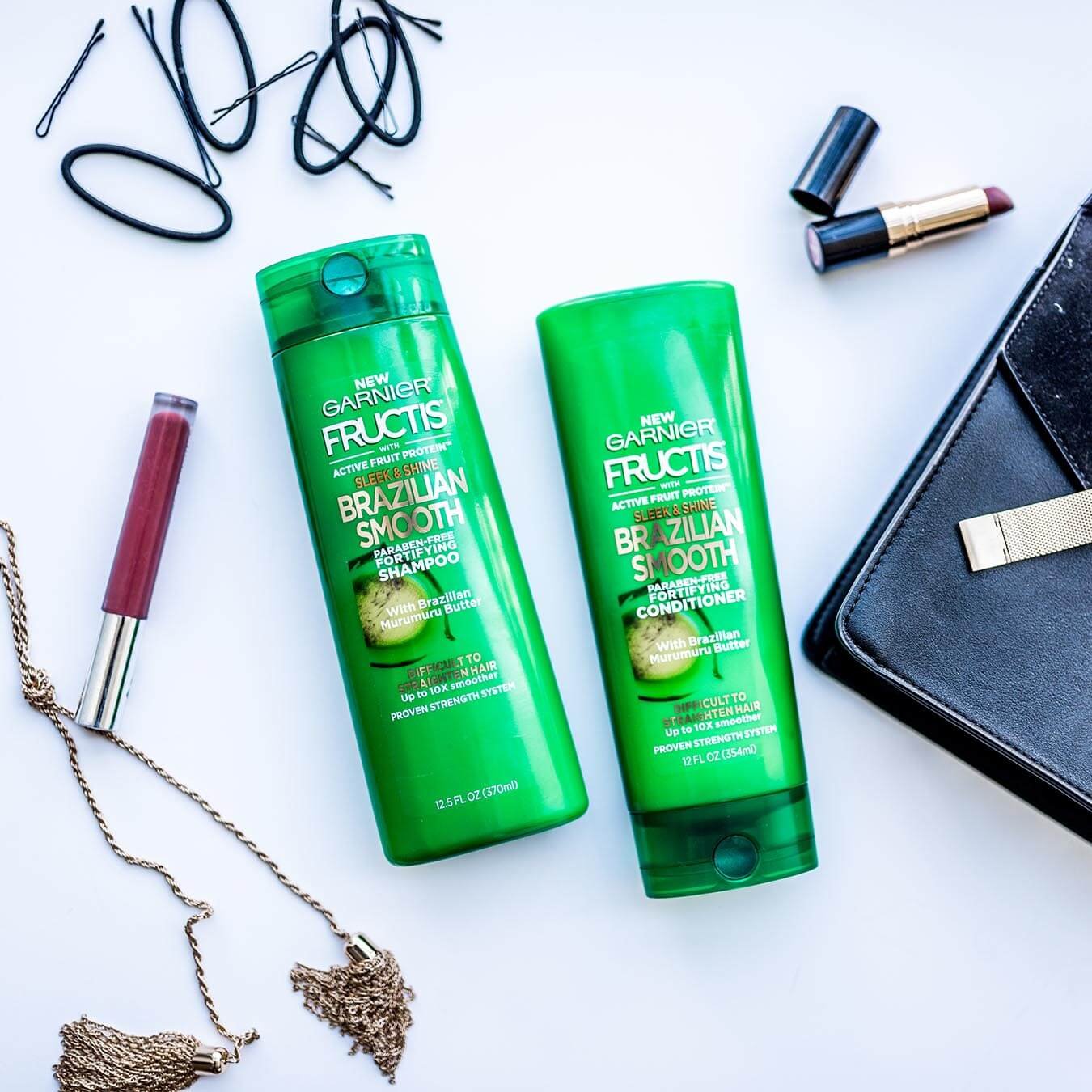 Garnier Fructis Brazilian Smooth Shampoo and Brazilian Smooth Conditioner on a blue background next to a leather purse, an open red lipstick, lip gloss, a knotted metal gold tassel, and several bobby pins and hair ties.