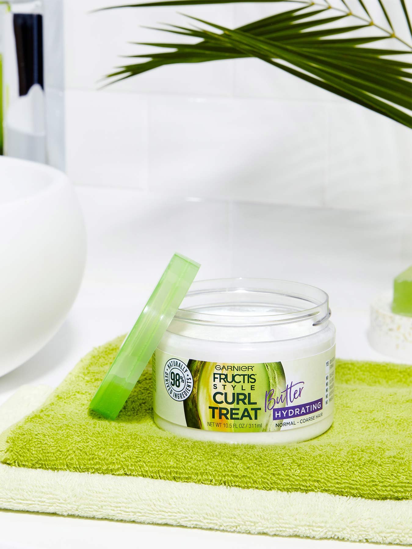 Open container of Curl Treat Butter Hydrating Leave-in Styler to Shape Curls on a green mat in a white bathroom with a plant.