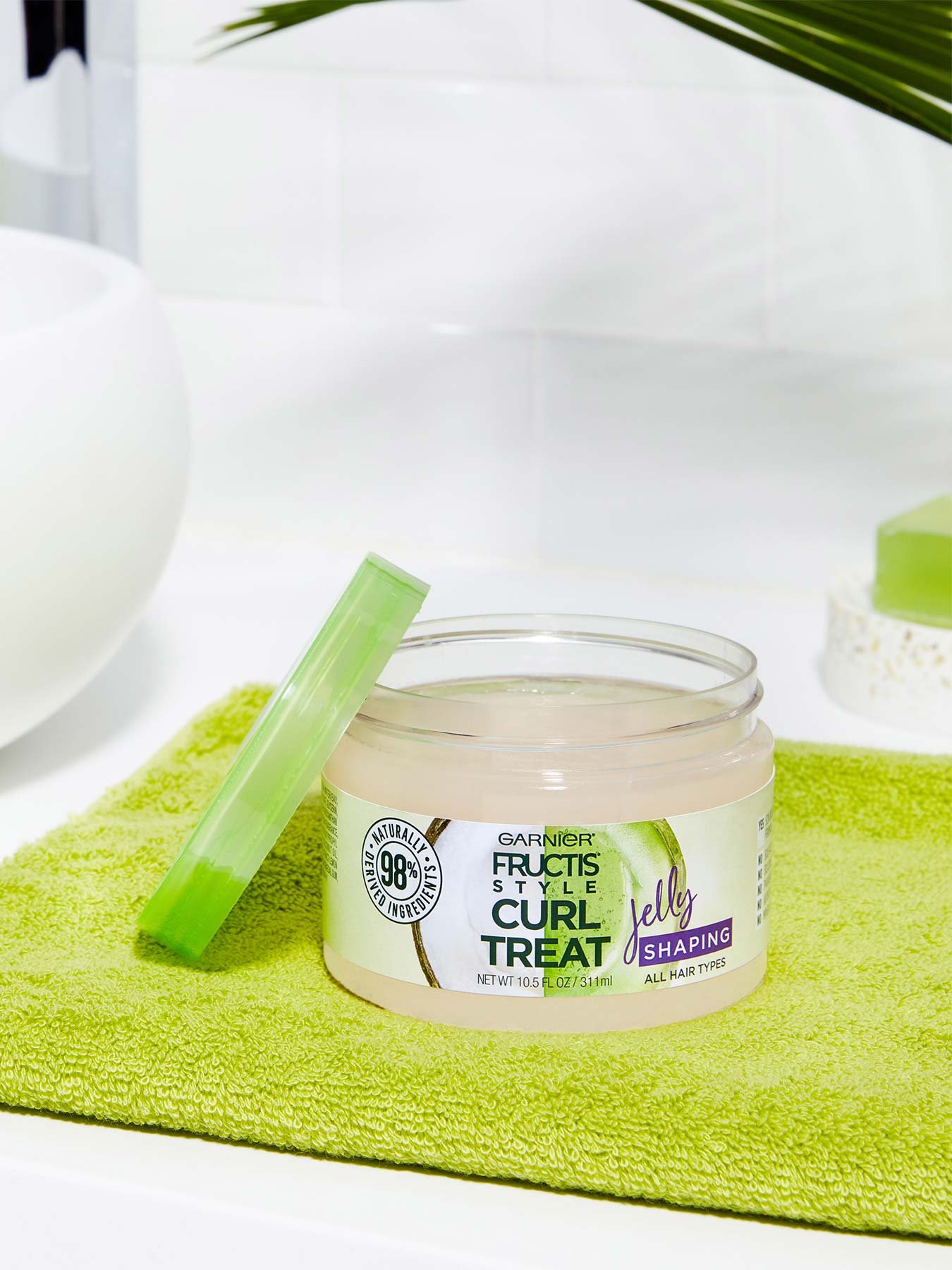 Open container of Curl Treat Jelly Shaping Leave-in Styler to Shape Curls on a green mat in a white bathroom with a plant.
