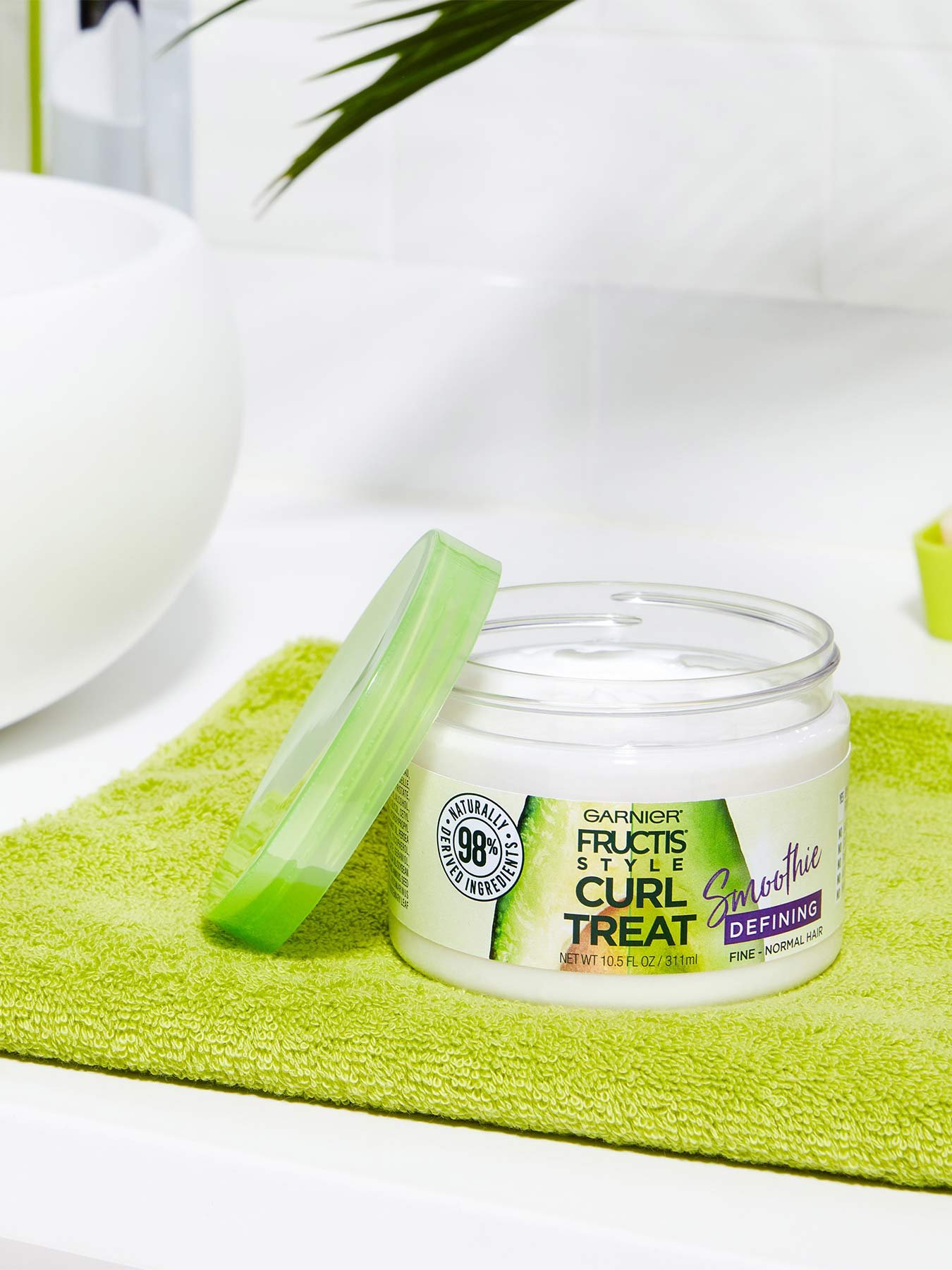 Open container of Curl Treat Smoothie Defining Leave-in Styler for Soft Curls on a green mat in a white bathroom with a plant.