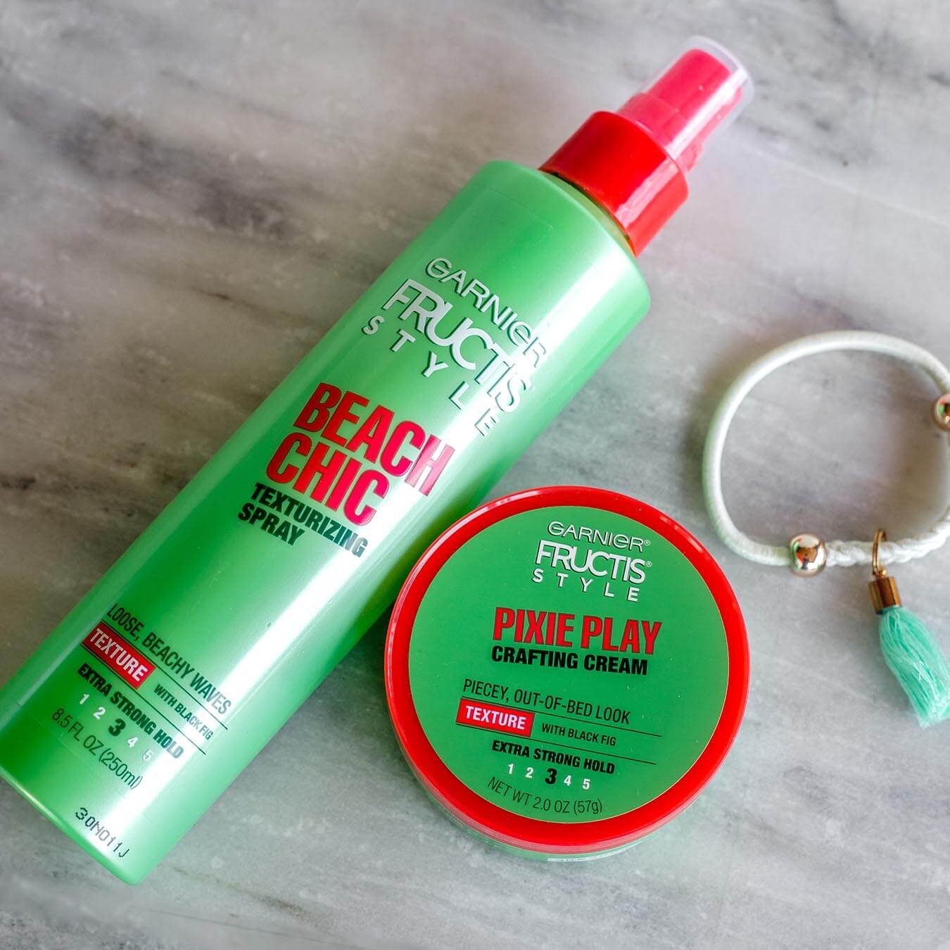 Garnier Fructis Style Beach Chic Texturizing Spray and Fructis Style Pixie Play Crafting Cream on gray marble next to a white bracelet with gold beads and a green tassel.