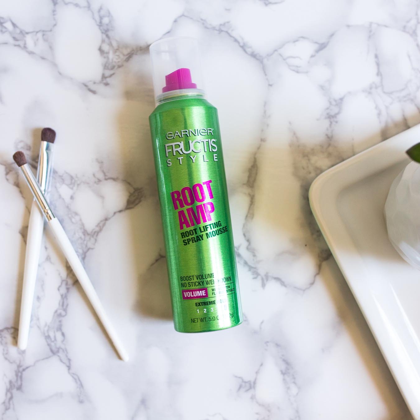 Garnier Fructis Style Root Amp Root Lifting Spray Mousse on white marble next to two makeup brushes and a white tray holding a succulent potted in a white faceted pot.